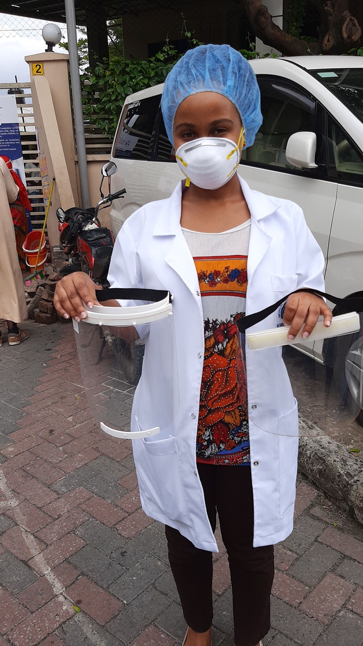 A woman wearing a lab coat, face mask and hair covering holds two plastic face shields