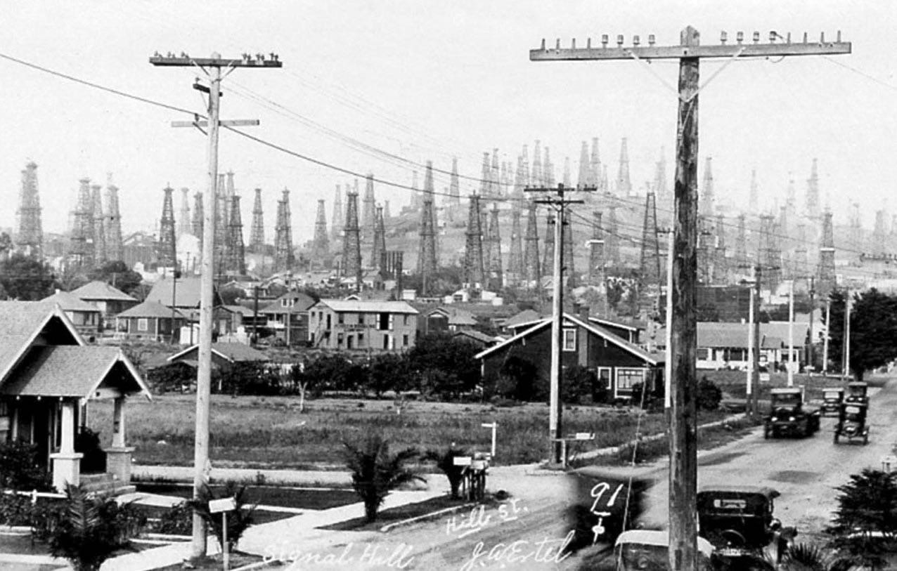 A historic black-and-white photo shows a street with houses, old cars and dozens of oil derricks on the hill behind them.