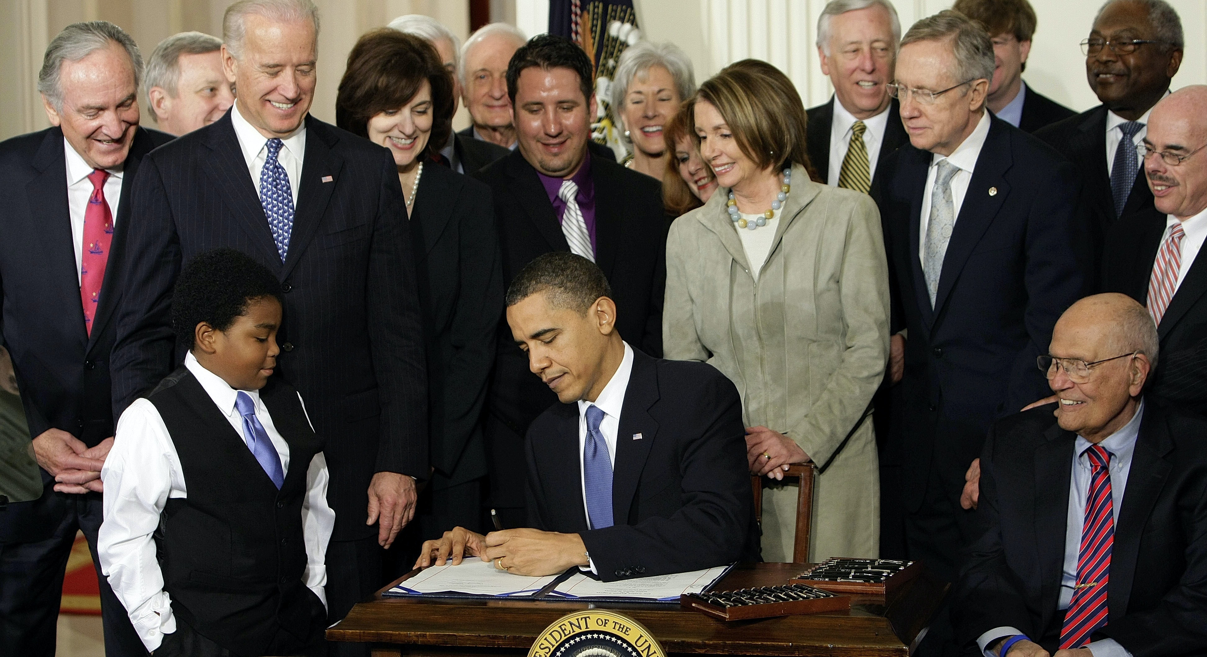President Barack Obama is surrounded by onlookers as he signs a document. Joe Biden, Nancy Pelosi and a young Black boy in a vest and tie are part of the crowd.