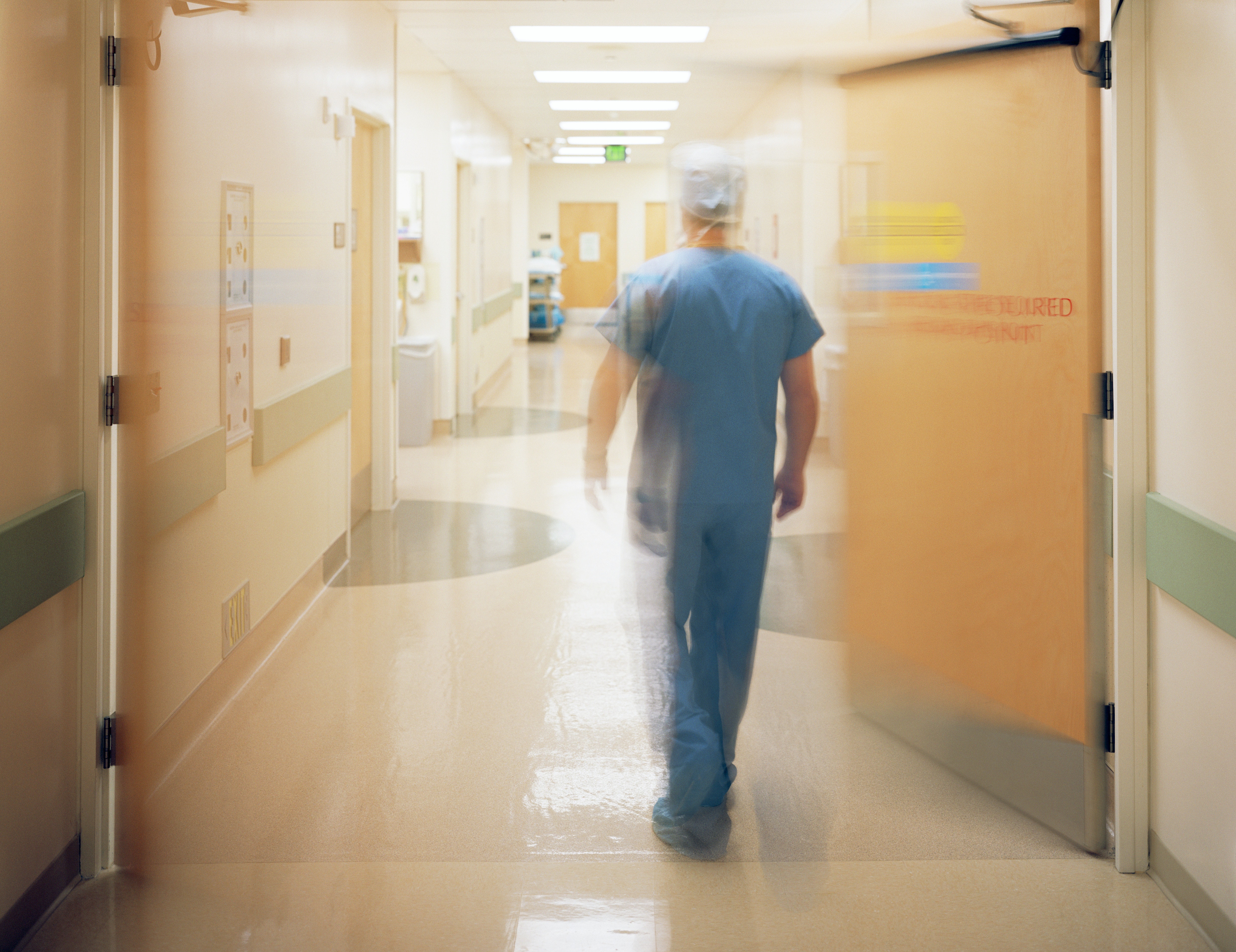 male surgeon in cap and blue scrubs walking down hospital hallway away from camera