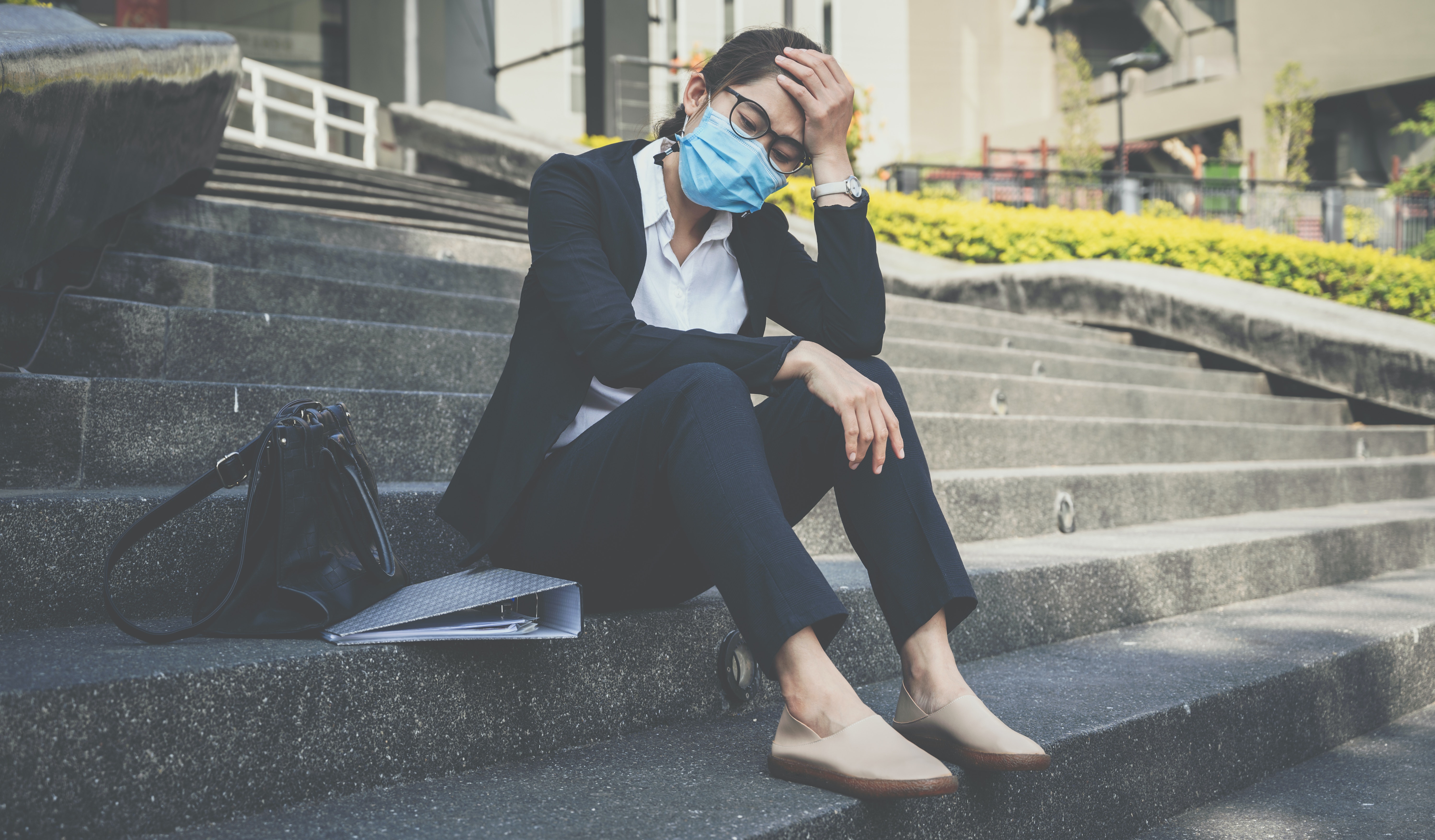 Woman wearing a surgical mask sits on the steps of an office building, resting her elbow on a bent knee holding her forehead, with her purse and work binder beside her.