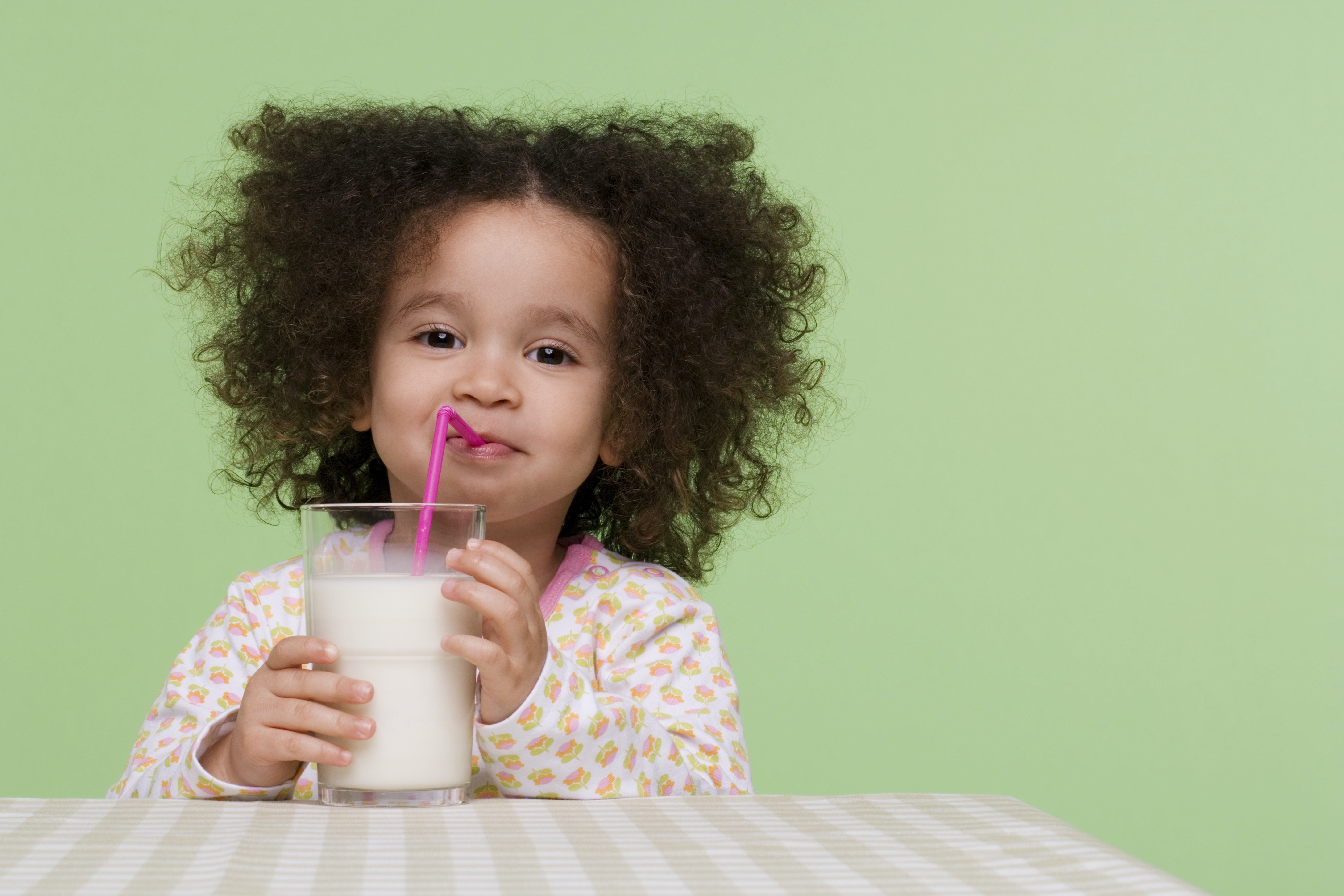 A smiling young girl drinking a glass of milk through a straw.