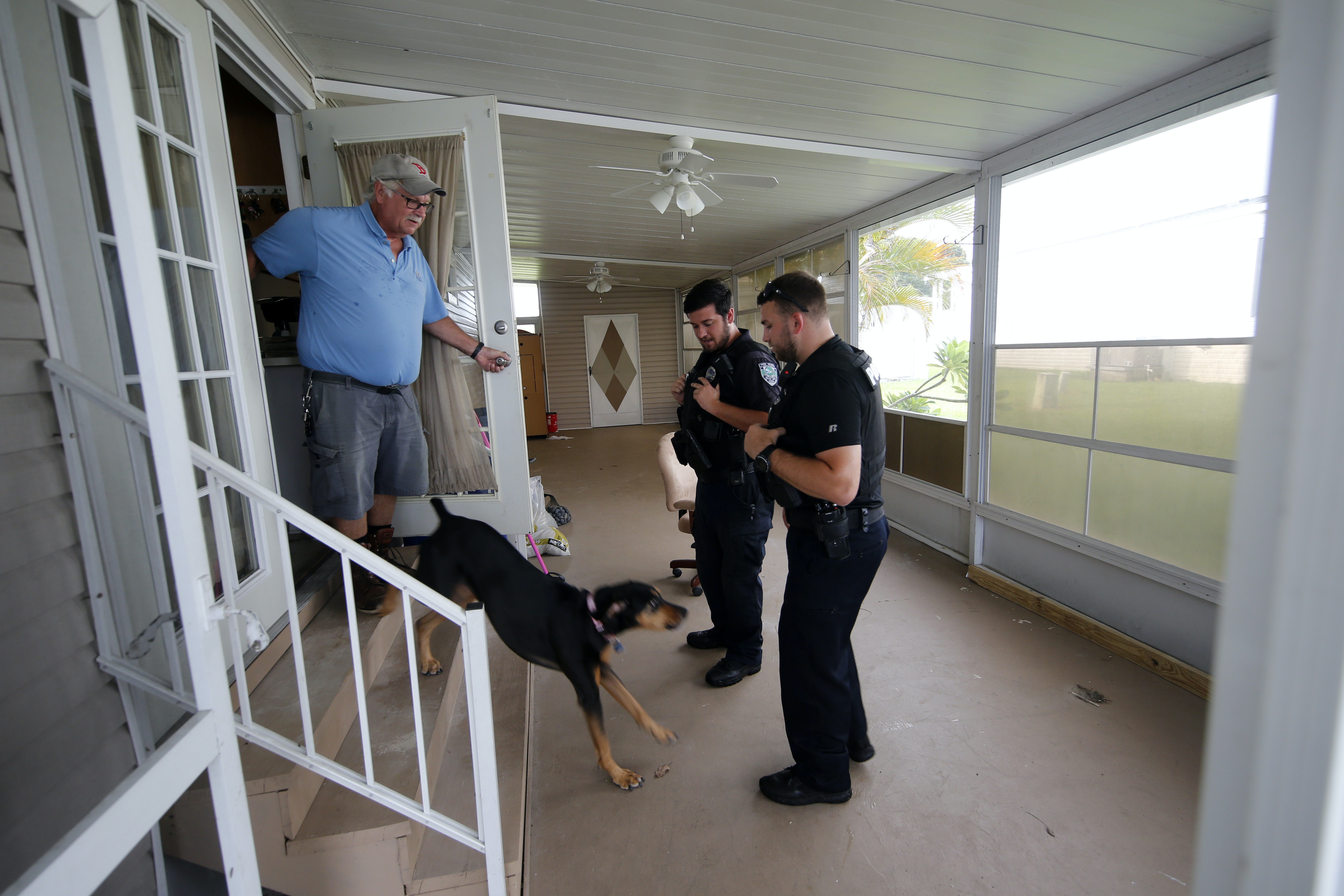 A man stands in the door of a mobile home as his dog runs out to greet two police officers.