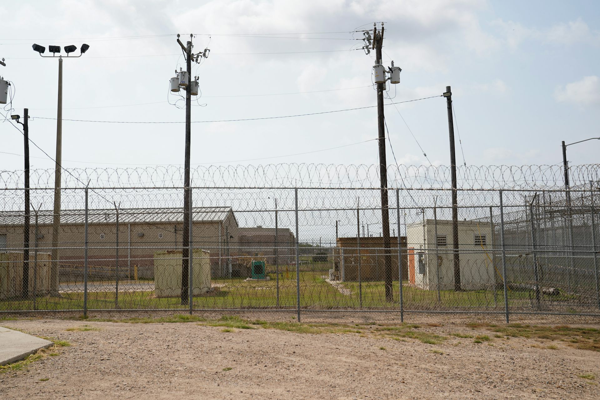 Detainee yard with low buildings behind fences topped with barbed wire and tall light poles