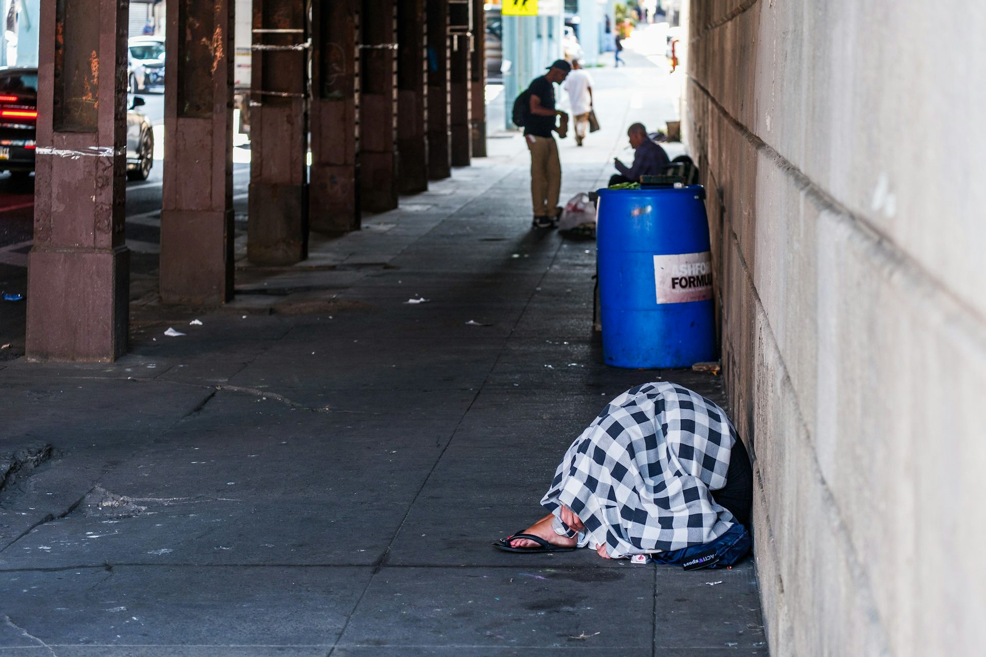 Under a bridge, a person crouches under a blanket in the foreground and two people talk in background