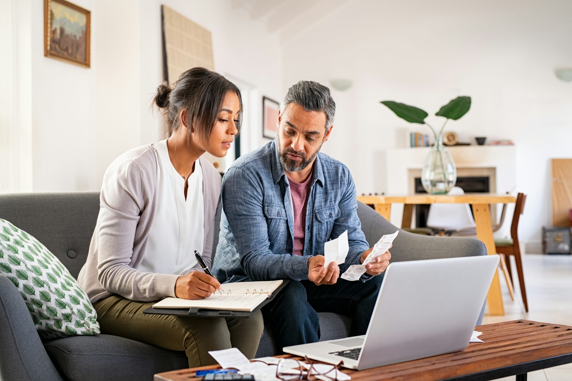 Middle-aged couple sits on couch with bills and planner in front of them, a laptop in the foreground.