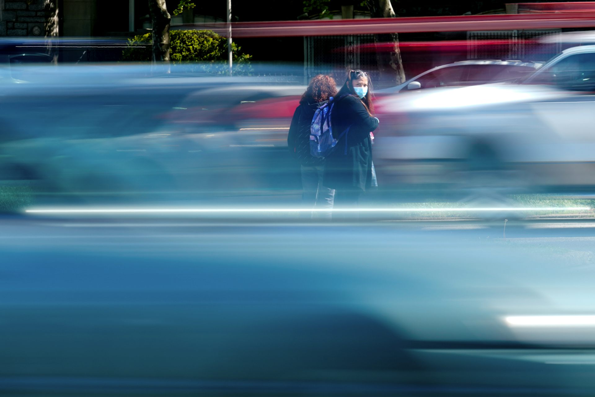 Two girls wearing school uniforms and backpacks stand between busy opposing lanes of traffic