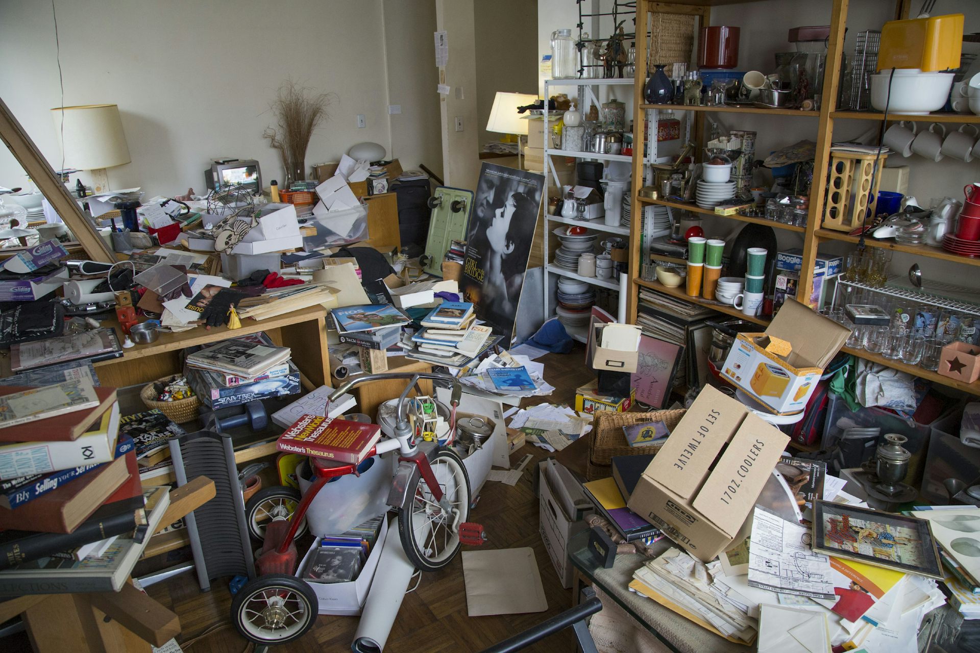 A room full of piles of papers, stacked shelves and a tricycle.