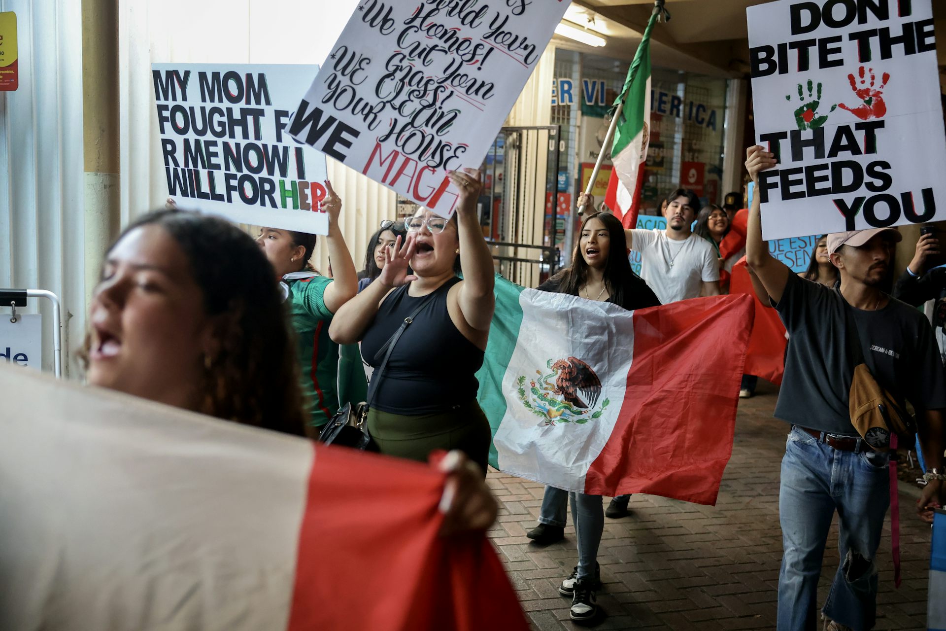 A group of protestors holding signs and yelling walk down the street. Some are holding Mexican flags.