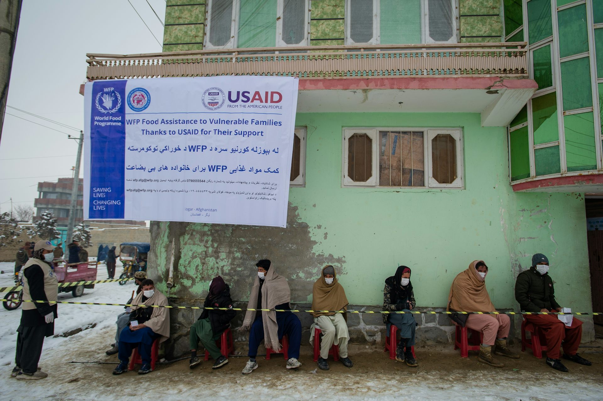 With a USAID banner hanging on a battered green building, a group of men sit outdoors, just below the banner.